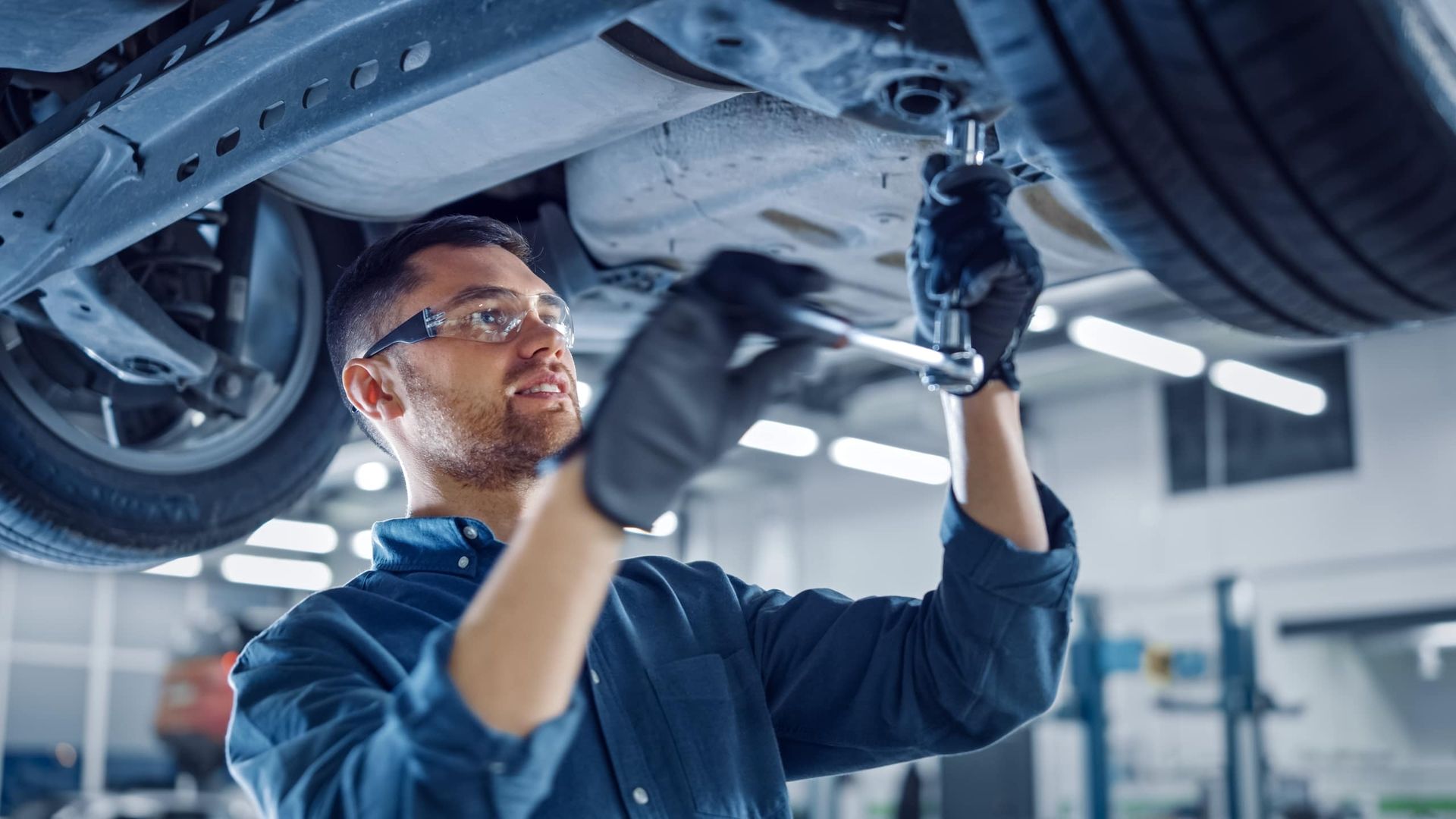 Kfz-Mechaniker mit Schutzbrille arbeitet mit einer Ratsche am Unterboden eines Fahrzeugs auf der Hebebühne